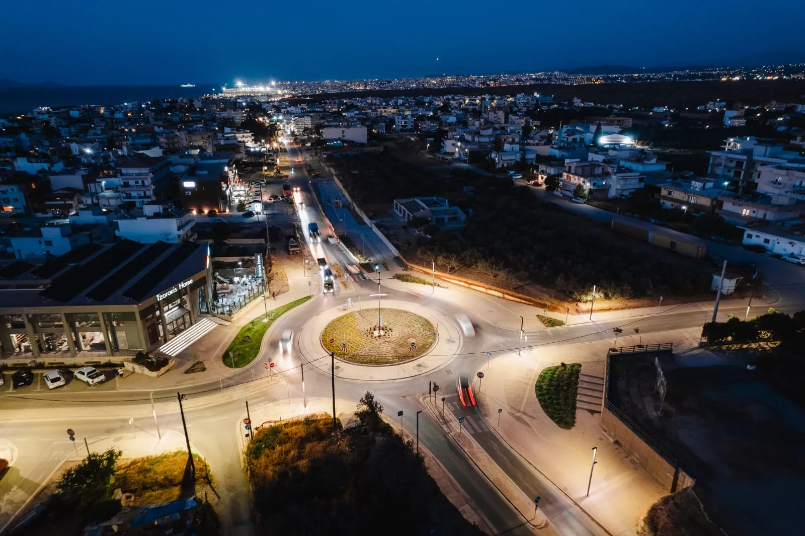 Night aerial of Gazi with the completed El Greco roundabout lit below, the town beyond and the sea on the horizon
