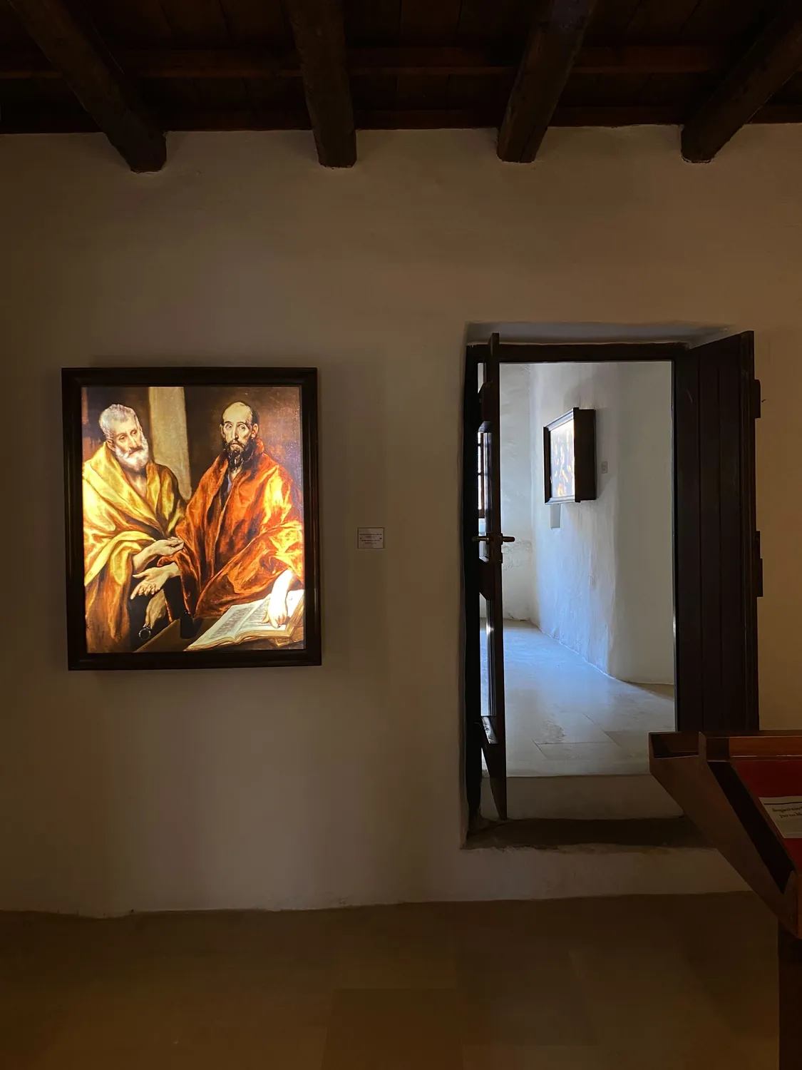 Interior of the Domenikos Theotokopoulos Exhibition Hall in Fodele, a framed El Greco work lit on a white wall beneath a timber-beamed ceiling