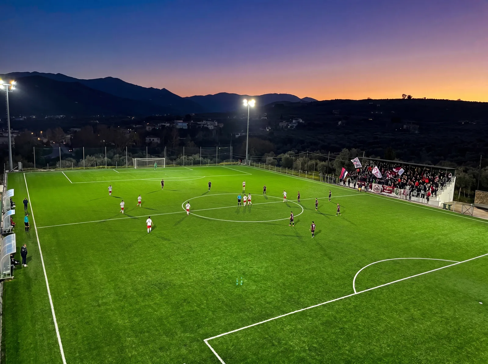 Night match at the Voukolies football ground with floodlights on and a full grandstand