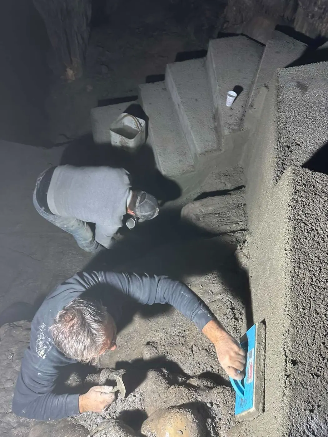 Two workers finishing a newly cast concrete step by worklight inside the Dikteon Cave