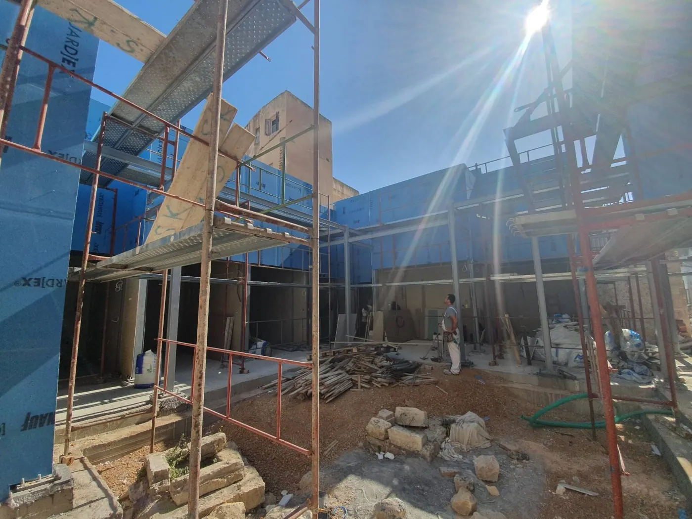 The steel frame erected and the shop units clad around the open courtyard in July 2021, with scaffolding and ancient stone blocks still on the courtyard floor
