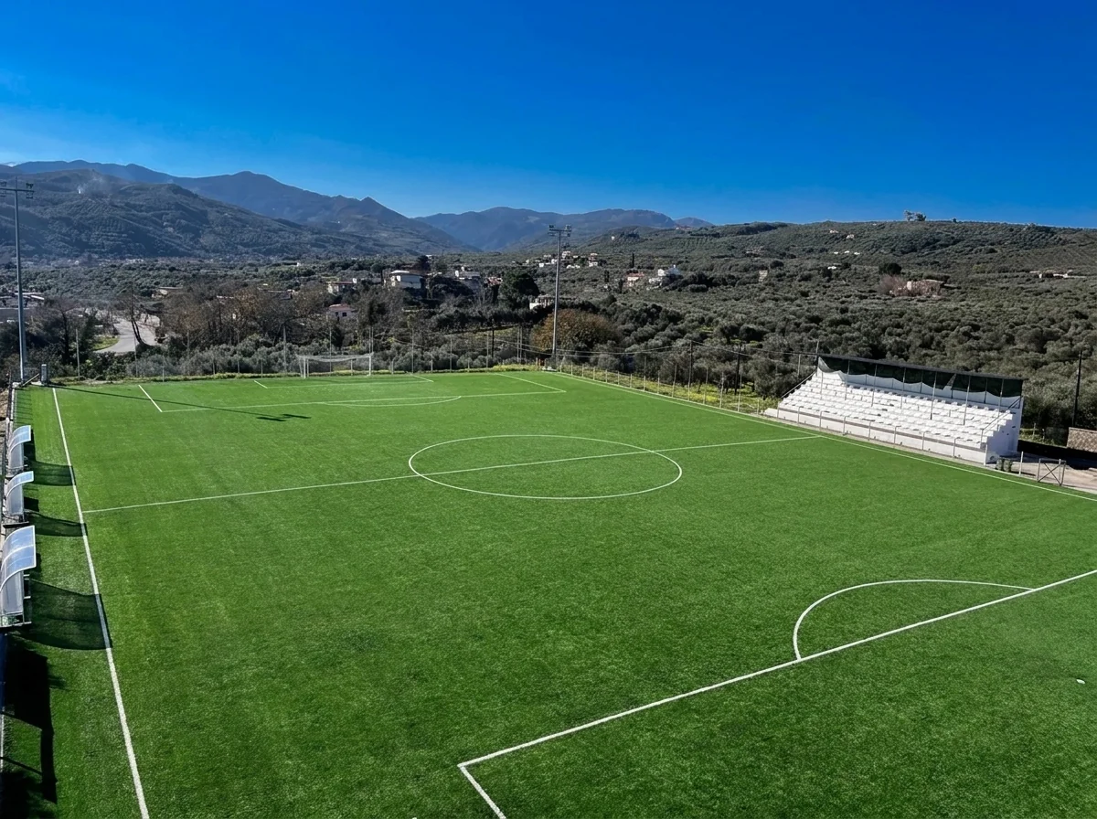 Aerial view of the Voukolies football ground in the olive hills of Chania — synthetic turf pitch, white grandstand and clubhouse, built by Lydakis Construction