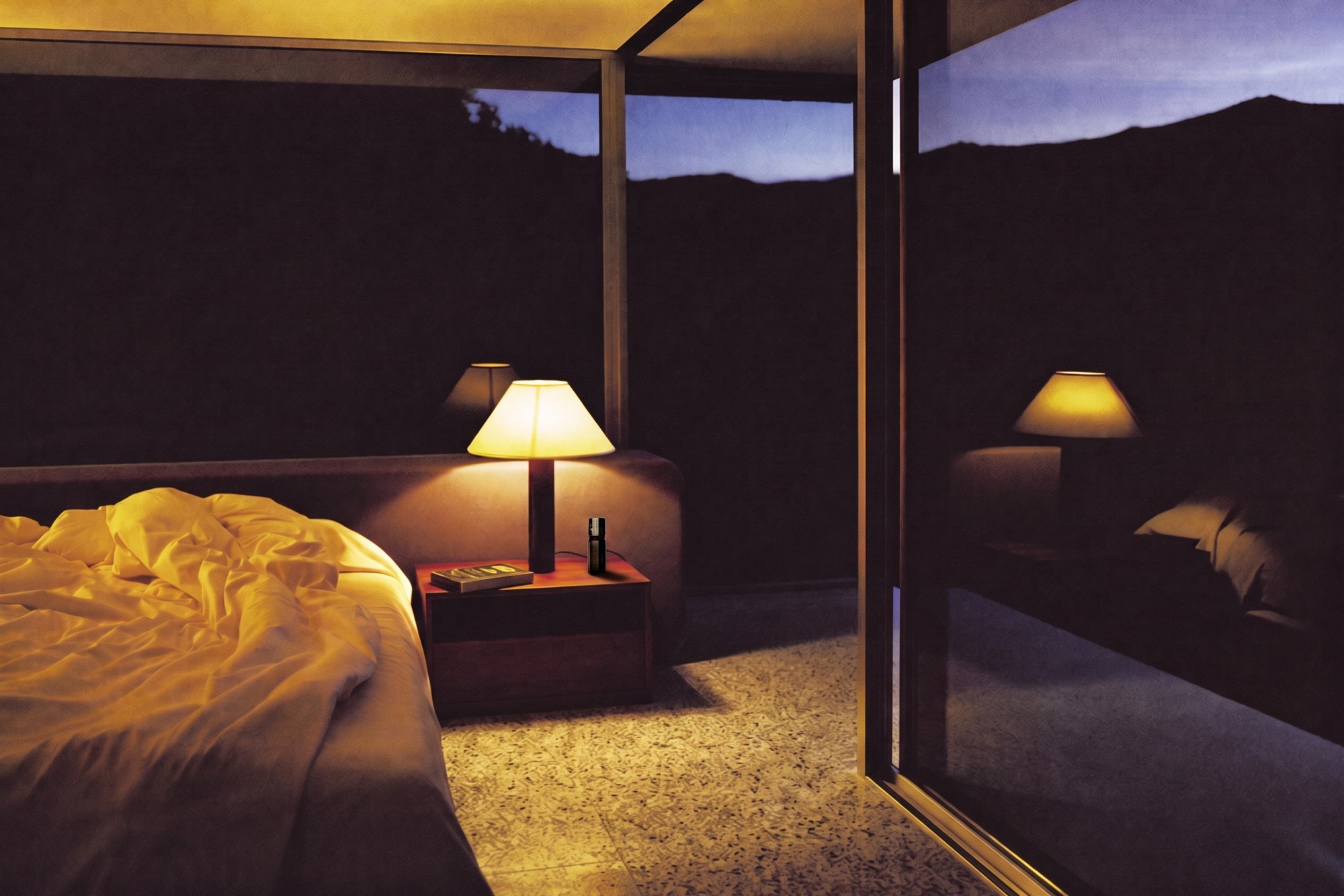 A mid-century modernist bedroom after dark, a single lamp on a teak block and black night beyond the glazing