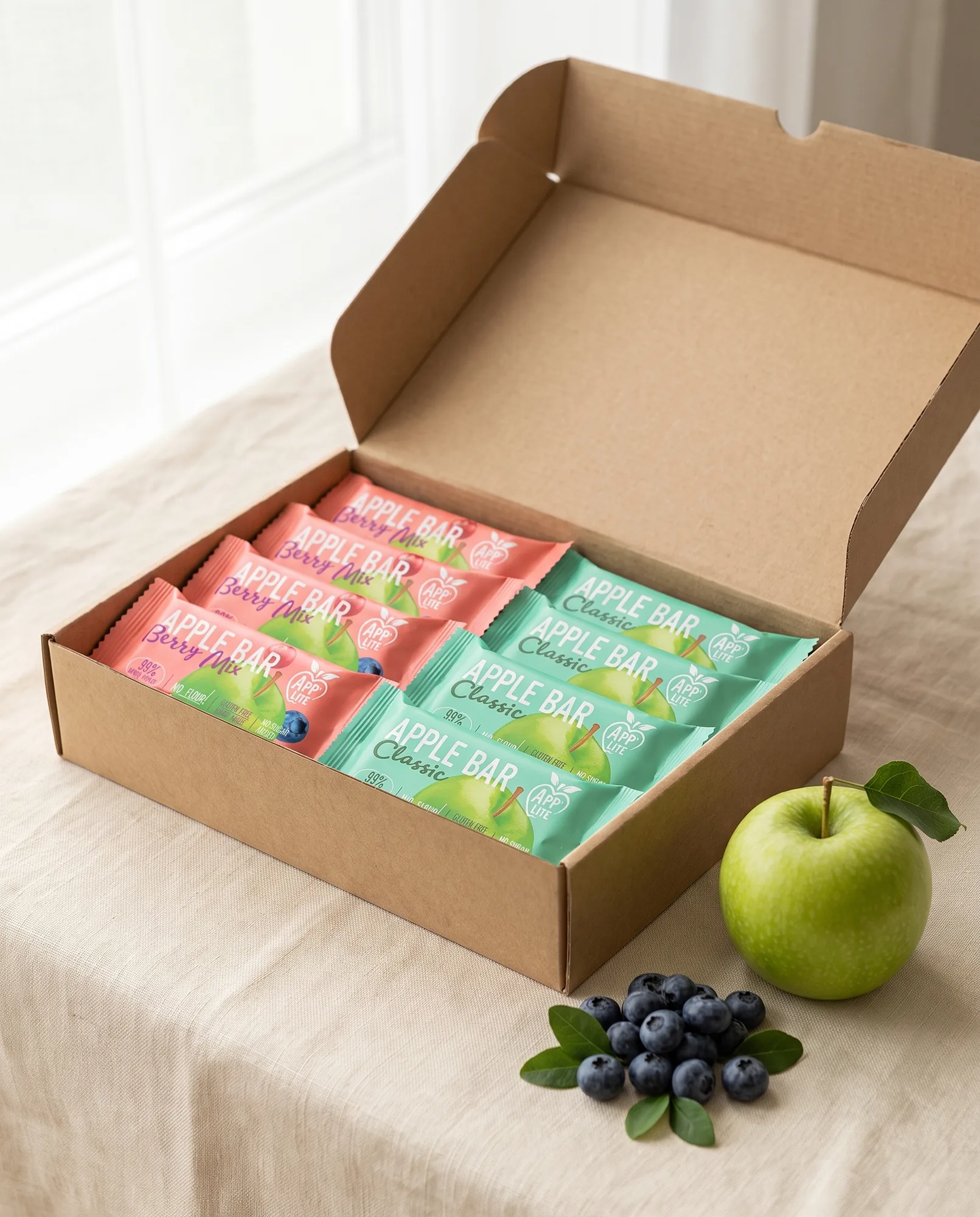 Open kraft box with twelve App'Lite Apple Bars in Classic and Berry Mix wrappers, a green apple and blueberries