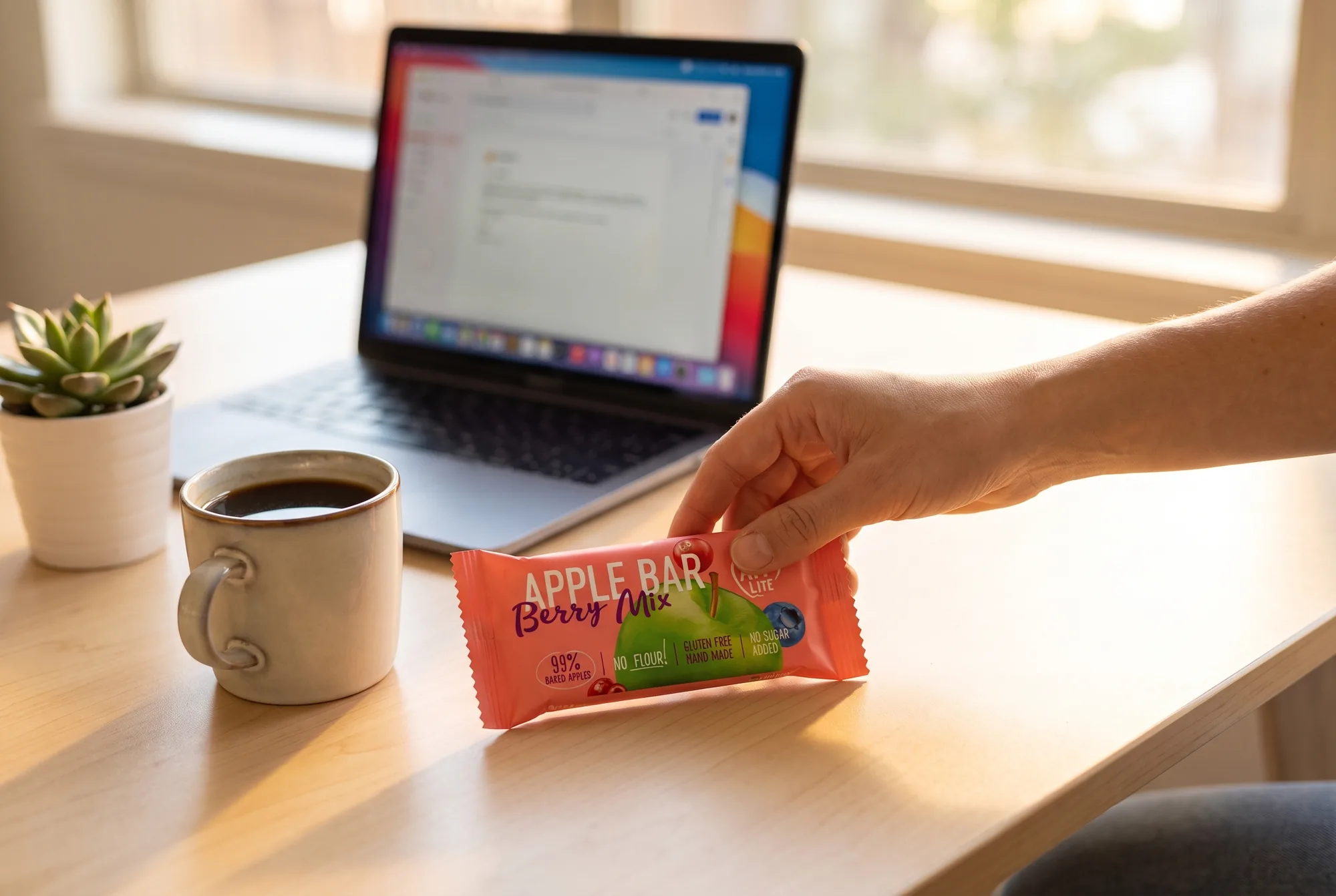 Hand reaching for a Berry Mix Apple Bar on a desk beside a laptop and coffee