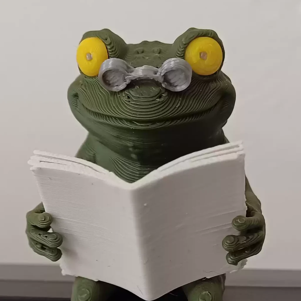 顔まわりの接写。鼻先にずり下がった丸メガネ、黄色い目、白い本、3Dプリントの積層痕。