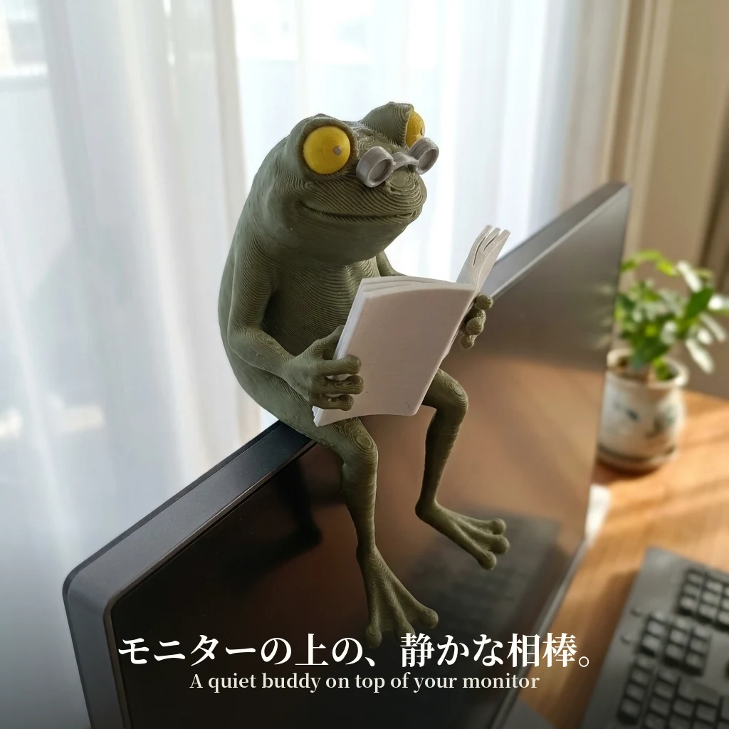 モニターの上のフチに腰かけ、後ろ足を垂らして読書するカエルの置物。