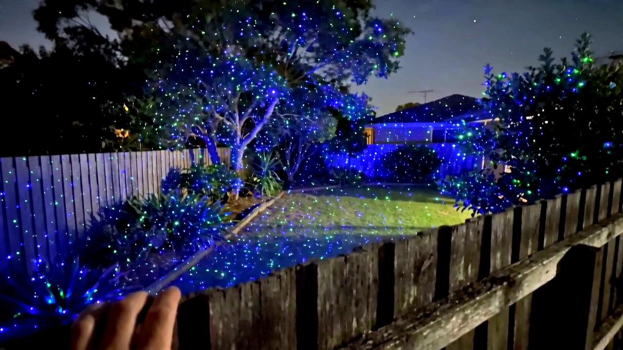 Frame from real customer phone footage showing green and blue laser stars covering a backyard, filmed over the fence at night