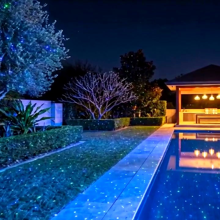 Still frame from real footage of green and blue laser stars across a poolside garden at night