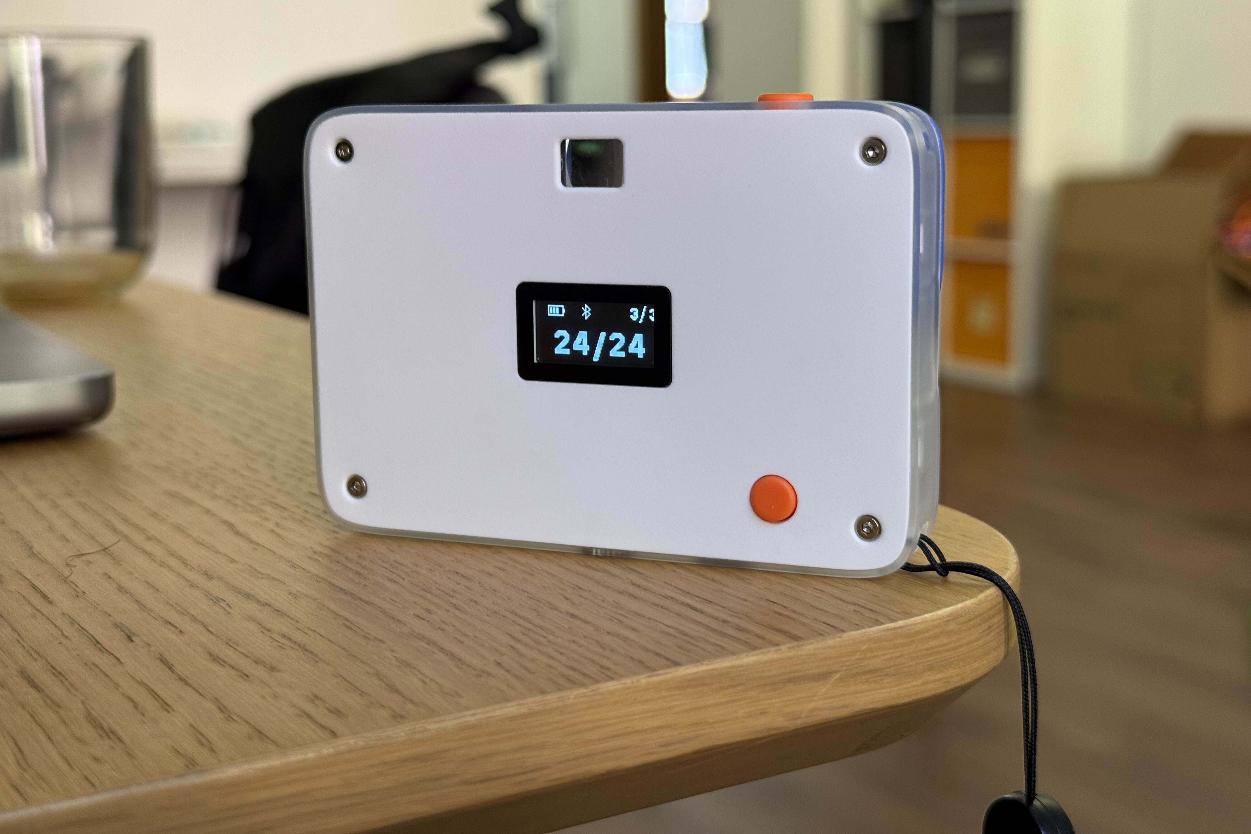 The white back of the Await camera with a small OLED counter showing 24 of 24, an orange shutter button and exposed Torx screws