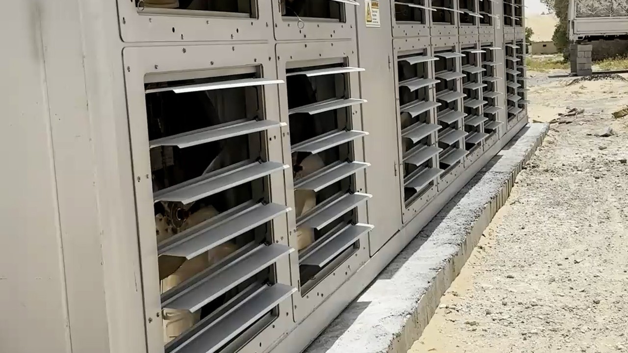 Row of louvred containerised mining units at a OneMiners hosting site in the UAE, desert ground and concrete apron in daylight
