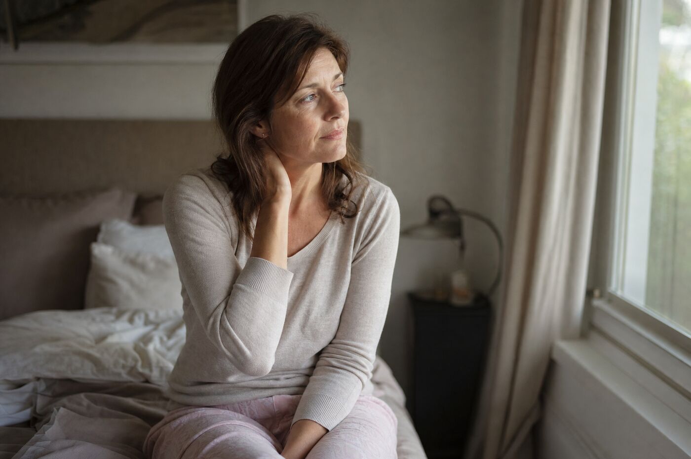 A woman in her forties sitting on the edge of her bed in the early morning, looking tired and thoughtful