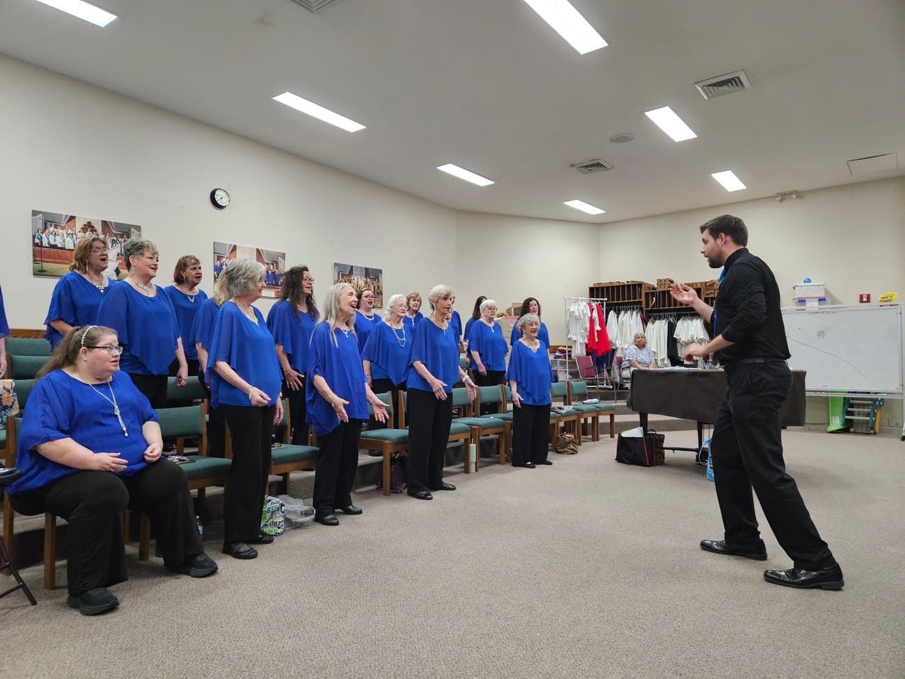 The chorus in blue tops singing in their rehearsal room while their director leads