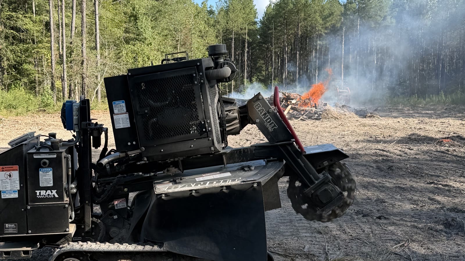 The Burton Stump Removal grinder on a cleared lot with the brush pile burning off against a pine treeline