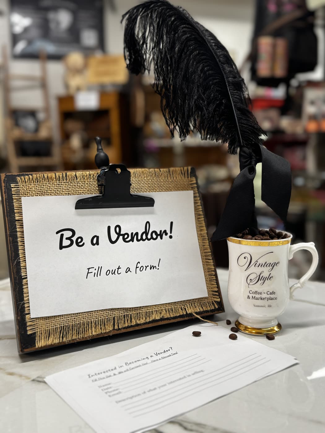 A &lsquo;Be a Vendor!&rsquo; sign on a burlap clipboard beside a Vintage Style coffee mug and a paper vendor form