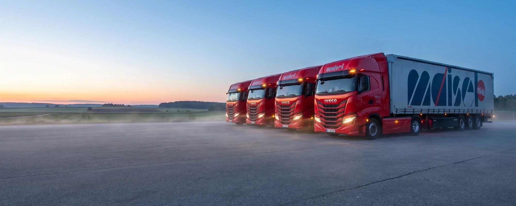 Vier rote IVECO S-Way Sattelzuege von Maisel in einer Reihe auf dem Hof in der Morgendaemmerung